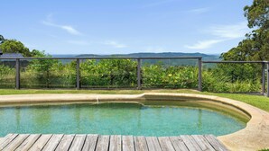 A swimming pool with a wooden deck, surrounded by lush greenery and a clear view of distant hills. - Hinterland Escape - Coastal Views, Pool & Fire Pit (Ninderry)