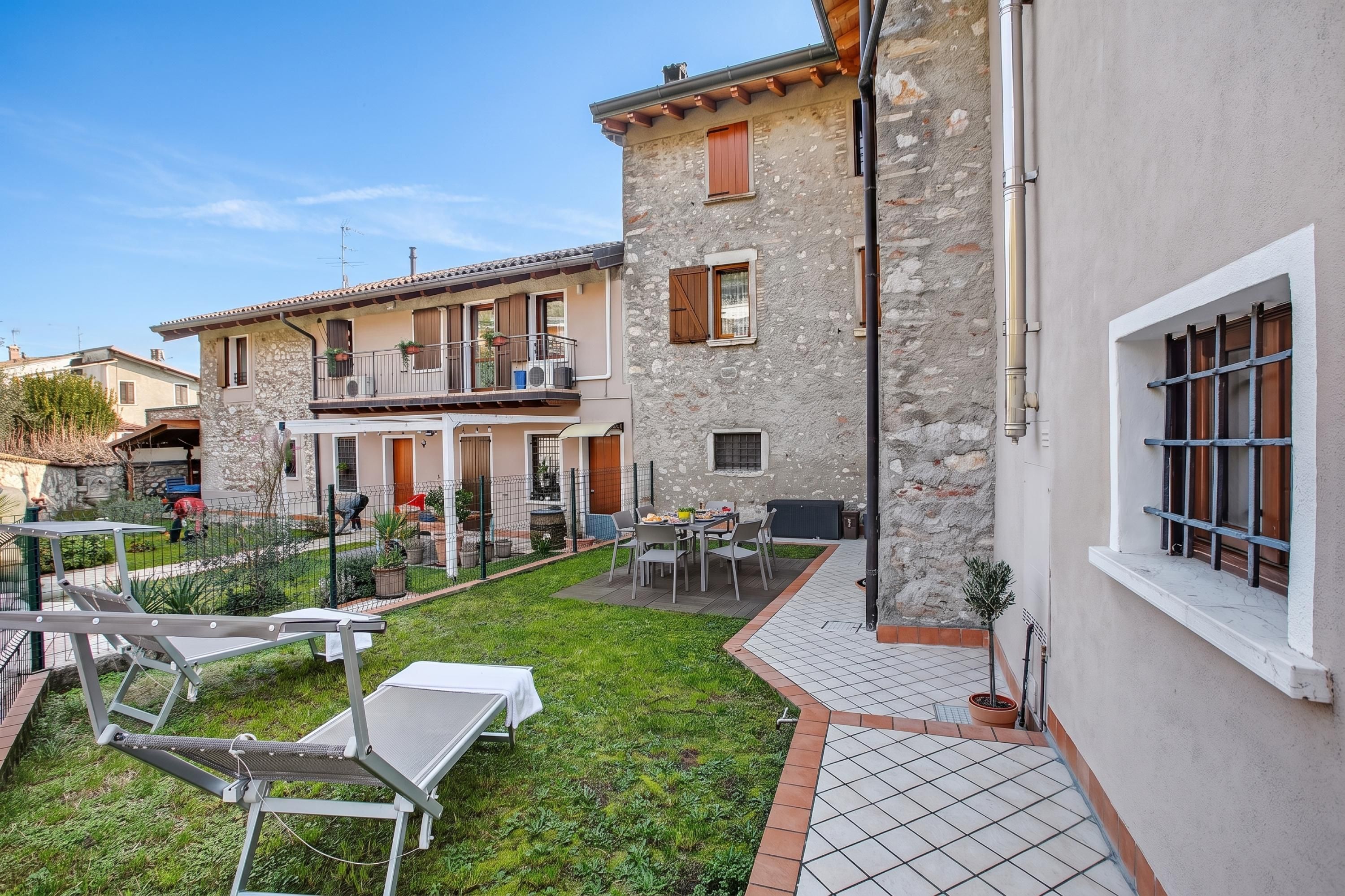 A courtyard with outdoor seating, a stone building with a balcony, and a tiled pathway.
