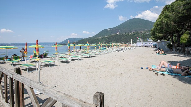 Une plage bordée de rangées de chaises longues et de parasols, une jetée en bois et, en arrière-plan, un paysage montagneux.