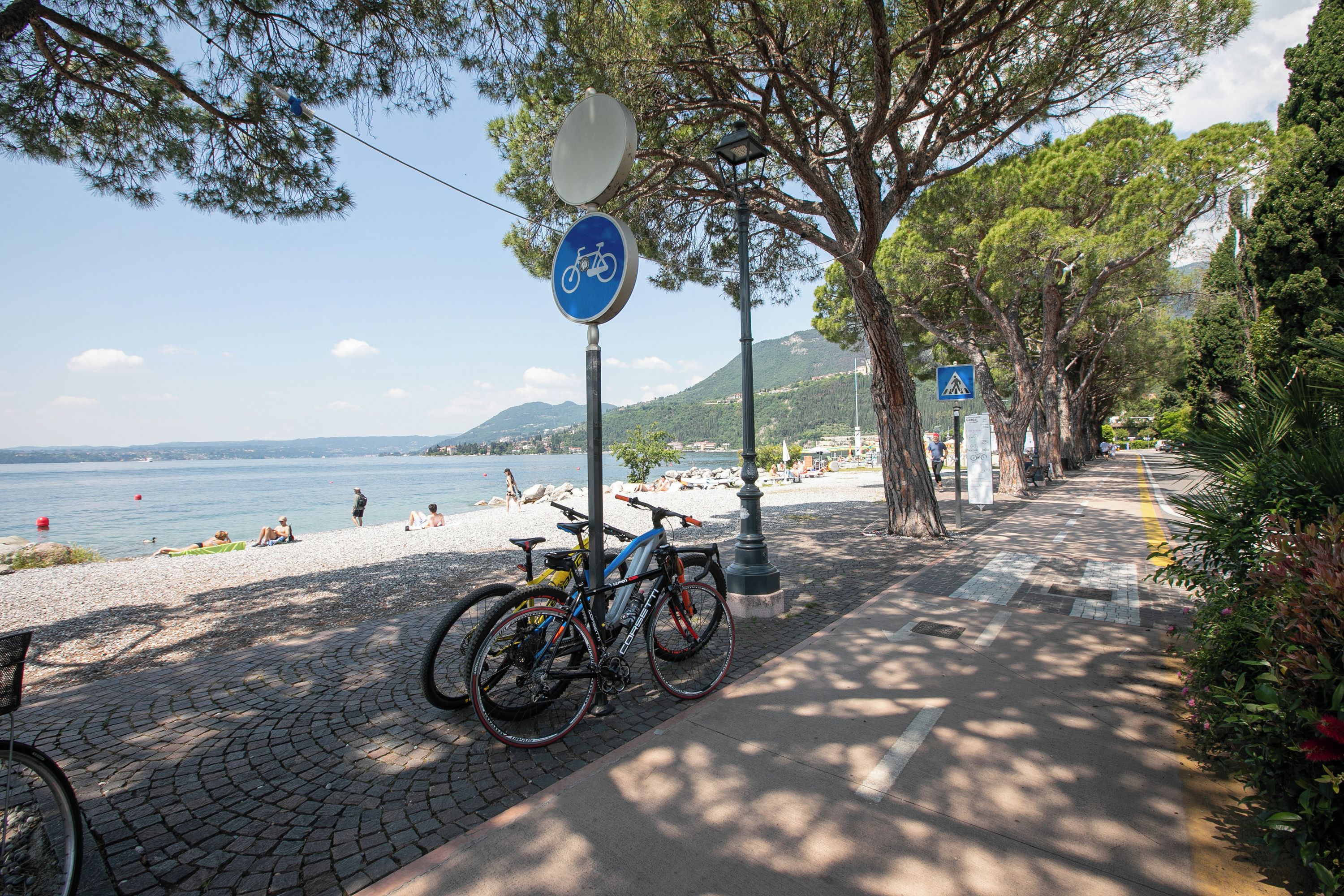 A paved pathway with bicycles parked, a beach in the background, and a clear sky.