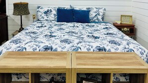 A bedroom with a bed, a bedside table, a lamp, a framed picture on the wall, and a shelf with a bottle and a sign. - Escape to the peaceful, Cottage 3:12 at Suttle Ridge in Arkadelphia, AR (Arkadelphia)