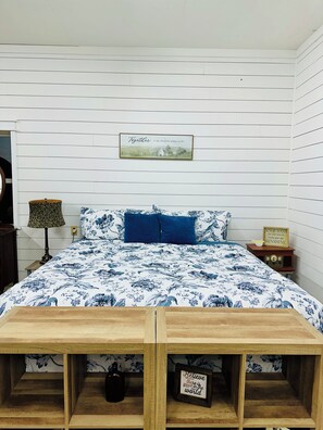 A bedroom with a bed, a bedside table, a lamp, a framed picture on the wall, and a shelf with a bottle and a sign.