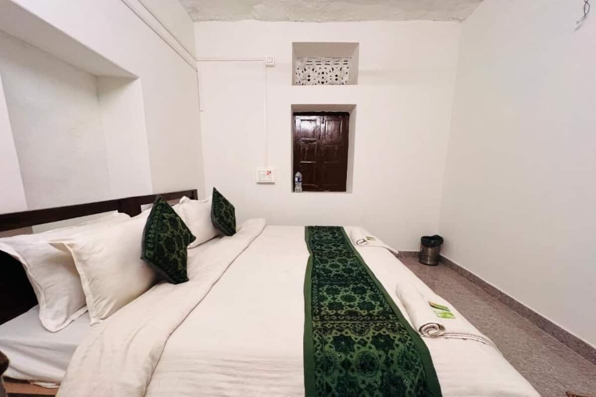 Een eenpersoonsbed met een wit-groene spreideken, een klein raam boven het bed, en een witte muur met een stopcontact.