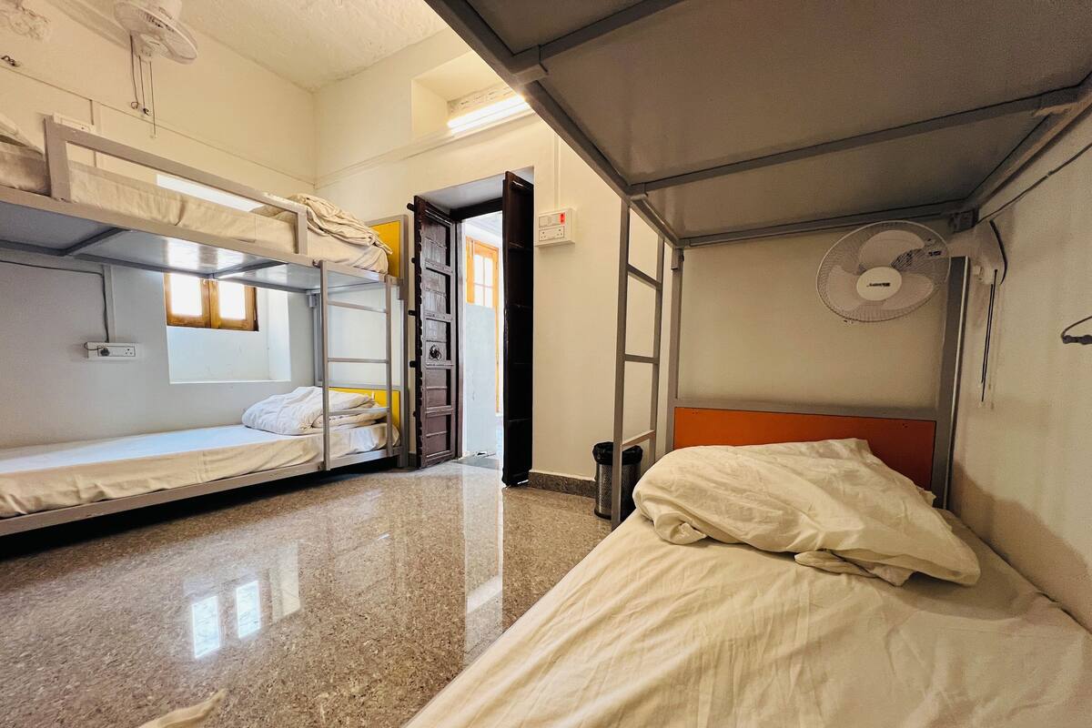 Een slaapzaal met stapelbedden, een ventilator en een afvalbak.