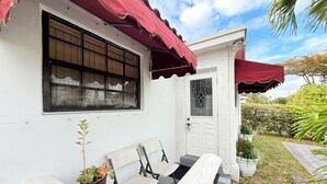 Een wit gebouw met een rode luifel, een raam met tralies, een deur met een ruit en een klein tuintje met stoelen en een potplant.
