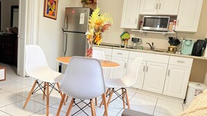 Een moderne keuken met witte kastjes, een roestvrijstalen koelkast, een magnetron en een koffiezetapparaat. Er staat een eettafel met vier stoelen bij, en op de tafel staat een vaas met bloemen.