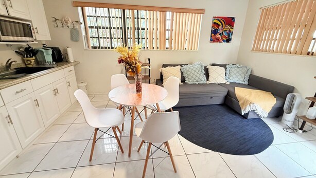 Een moderne keuken met witte kasten, een magnetron en een spoelbak. Er is een eethoek met een ronde tafel en witte stoelen. Een grijze bank en een zwart tapijt bevinden zich in de woonkamer.