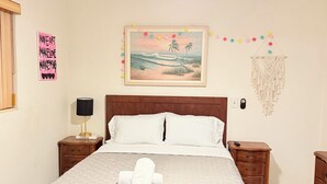 Een opgemaakt bed met een witte deken en een opgerolde handdoek, een plafondventilator, een schilderij van een strand, een wanddecoratie met een motiverende tekst, en een macramé wandkleed.