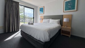 Ein Hotelzimmer mit einem großen Bett, einem Nachttisch mit Lampe, einem Fenster mit Vorhängen und zwei gerahmten Bildern an der Wand.