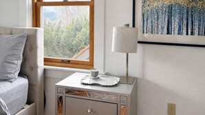 Un dormitorio con cama, una mesita de noche con lámpara, una obra de arte enmarcada y una ventana con vistas a árboles y una línea eléctrica.
