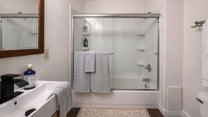 Un baño moderno con ducha envuelta en vidrio, un lavamanos blanco con dispensador de jabón y un perchero con dos toallas.