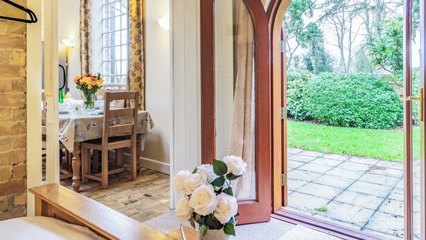 A room with a wooden bench, a dining table with chairs, a vase of flowers, and a view of a garden through an arched doorway. - Stearn Cottage-17917 (Bury St. Edmunds)