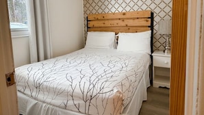 A bedroom with a wooden headboard, a bed with a leaf-patterned bedspread, a nightstand, and a window with curtains. - Whispering Pines Resort (Grand Bend)