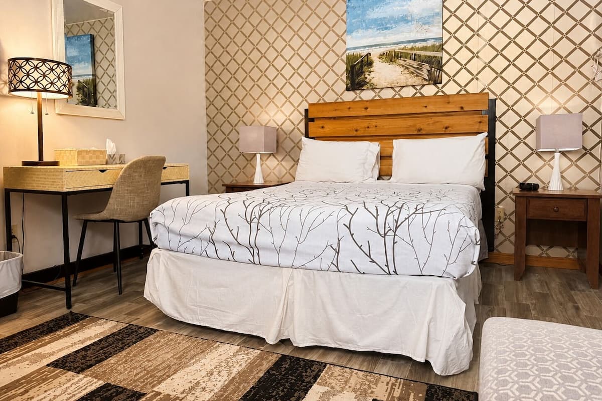A bedroom with a bed, two bedside tables, a desk, a chair, a lamp, a mirror, and a patterned wall.