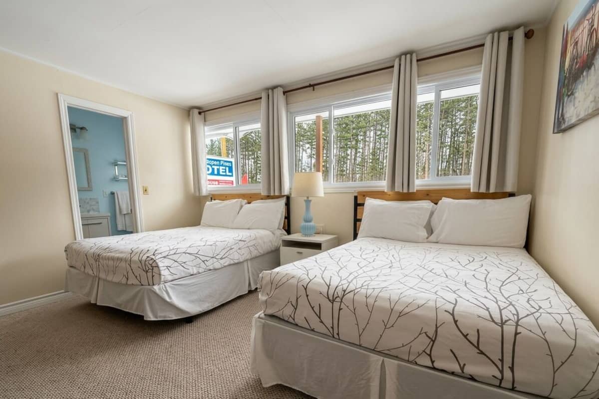 A hotel room with two beds, a nightstand, a lamp, and a view of trees through the window.
