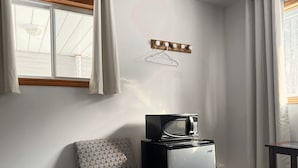 A compact room with a patterned chair, a small refrigerator, a microwave, a wooden shelf with hooks, and a window with curtains. - Whispering Pines Resort (Grand Bend)