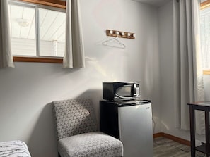 A compact room with a patterned chair, a small refrigerator, a microwave, a wooden shelf with hooks, and a window with curtains. - Whispering Pines Resort (Grand Bend)