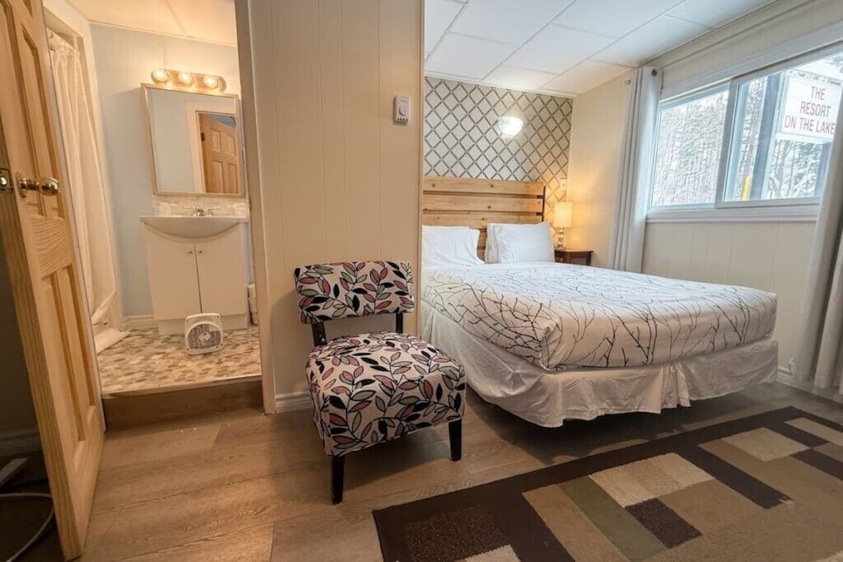 A bedroom with a bed, a chair, a vanity with a mirror, and a window with a view of trees.