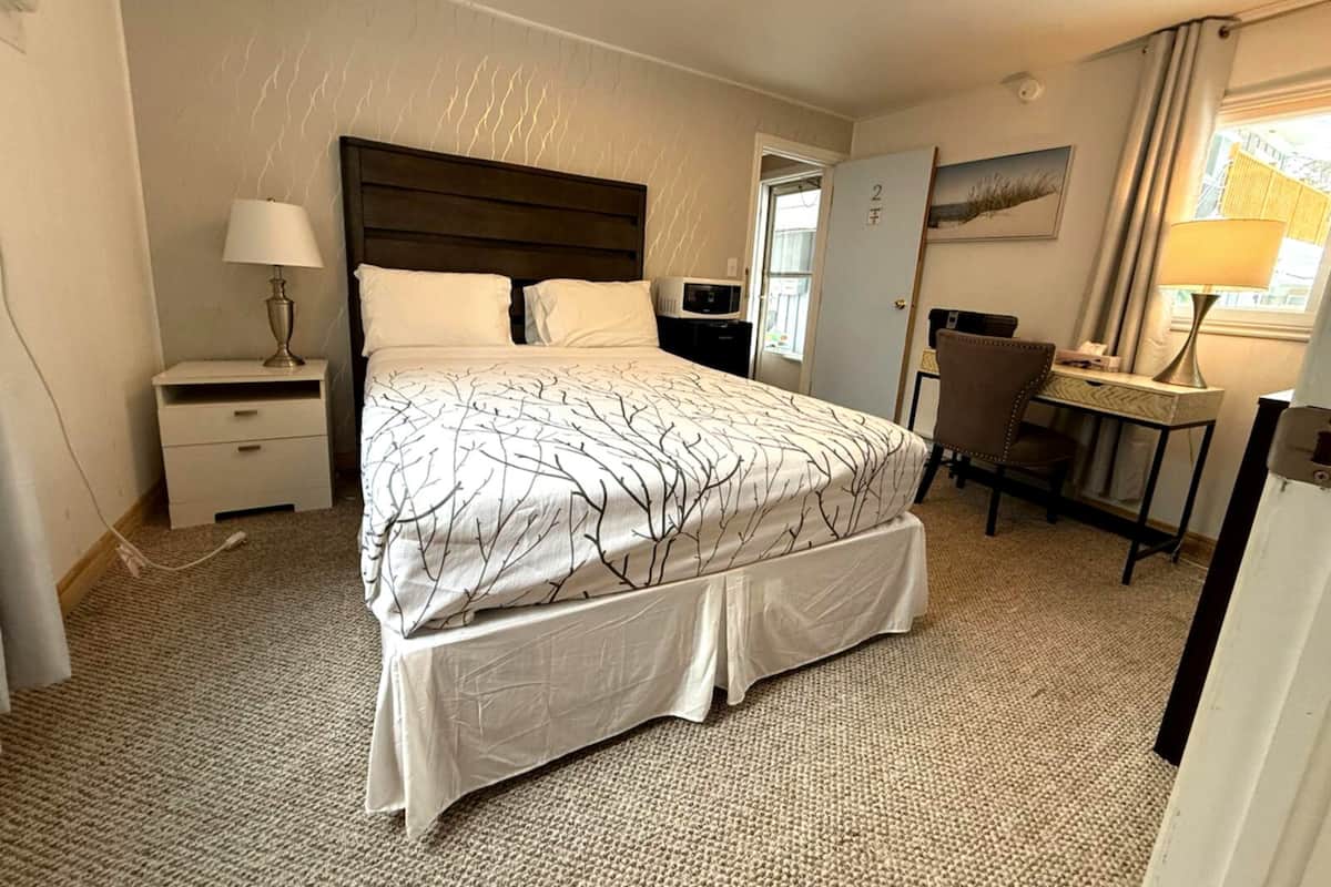 A hotel room with a bed, bedside table, lamp, desk, chair, and a door.
