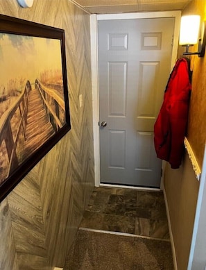 Un pasillo con piso de madera, una puerta gris, un cuadro enmarcado en la pared y una chaqueta roja colgada en la pared.