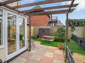 A patio area with a wooden pergola, a picnic table, and a glass door leading to a room with wicker furniture. - Anchors Away - 29566 (Blue Anchor, near Minehead)