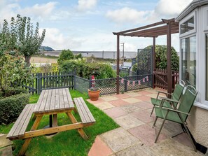 A patio with a wooden table and chairs, surrounded by a garden and a view of the sea. - Anchors Away - 29566 (Blue Anchor, near Minehead)