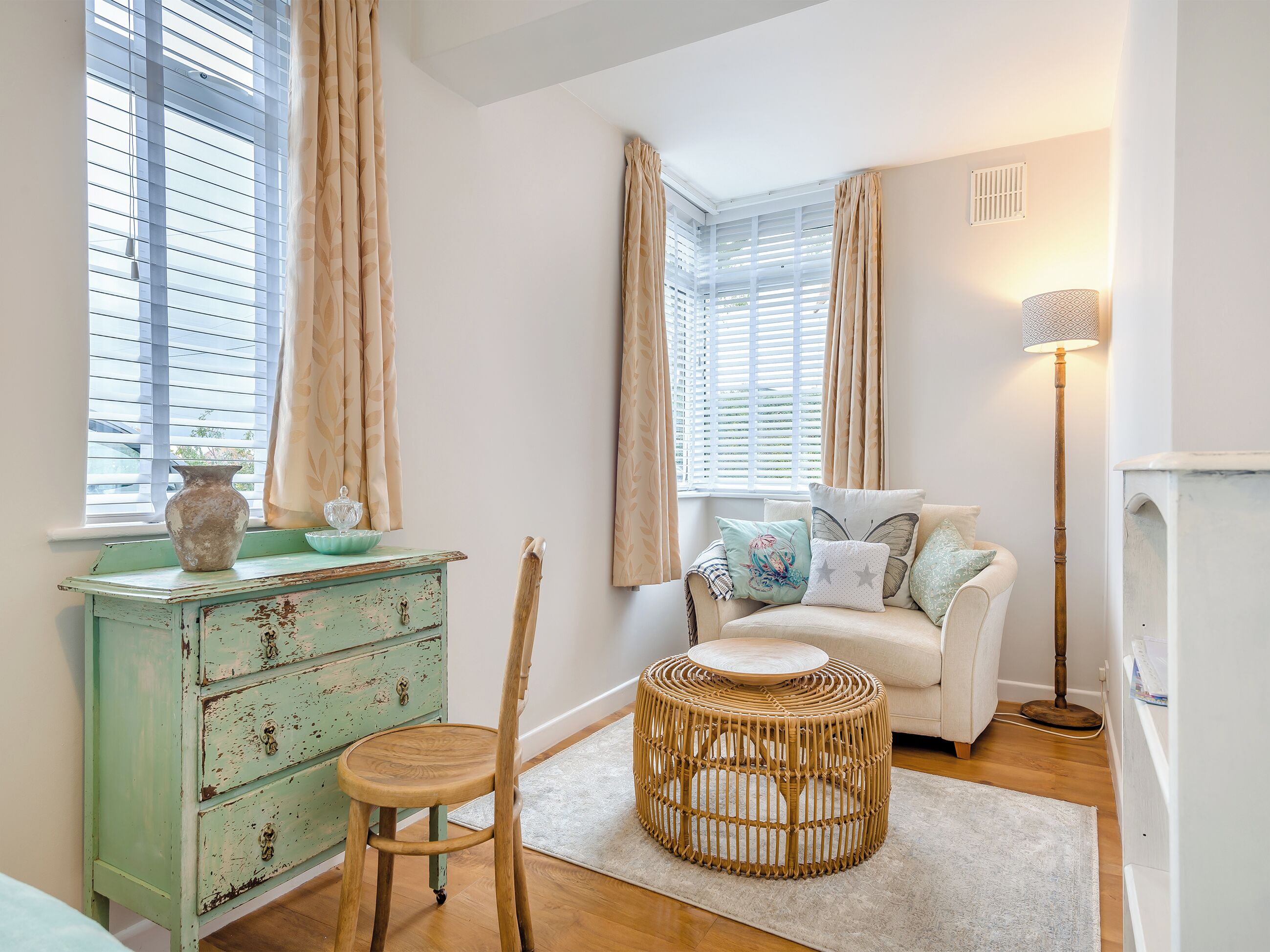 Une pièce chaleureuse avec une commode verte d’époque, un fauteuil en osier, un canapé blanc agrémenté de coussins à motifs et un lampadaire.