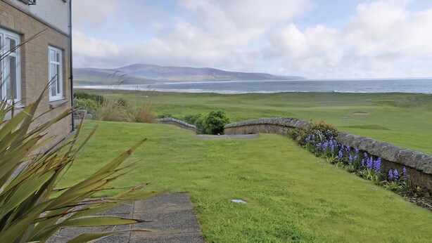 A coastal view with a grassy area, a stone wall with flowers, and a building with windows. - Links Apartment 8 - 29971 (Brora)