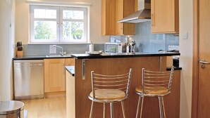 A modern kitchen with wooden cabinets, a stainless steel dishwasher, a microwave, and two bar stools. - Links Apartment 8 - 29971 (Brora)