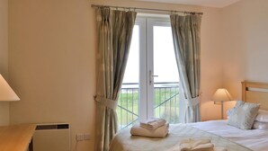 A hotel room with a bed, two bedside lamps, a window with curtains, and a view of the outdoors. - Links Apartment 8 - 29971 (Brora)