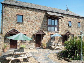 A stone building with a patio area featuring wooden benches and umbrellas, a wooden door, and a small window. - 2 bedroom accommodation in Marhamchurch, near Bude (Marhamchurch, near Bude)