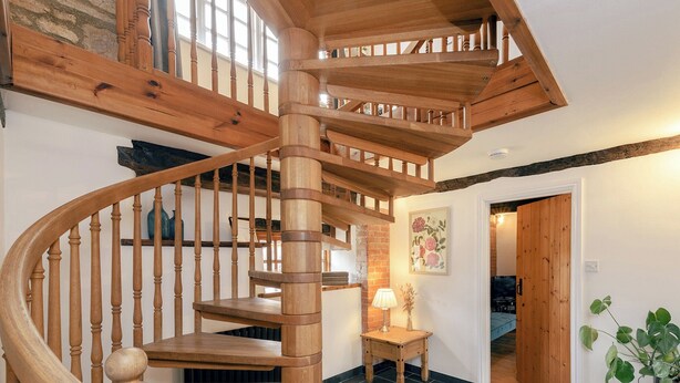 Una escalera espiral de madera en una habitación con suelo de baldosas, una pequeña mesa con una lámpara y una puerta que da a otra habitación.