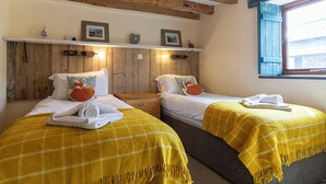 Een kamer met twee bedden, houten balken in het plafond, een raam met blauwe luiken en een plank met ingelijste foto's.