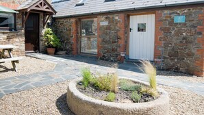 A stone building with a wooden door, a small window, and a patio area with plants and a table. - 2 bedroom accommodation in Marhamchurch, near Bude (Marhamchurch, near Bude)