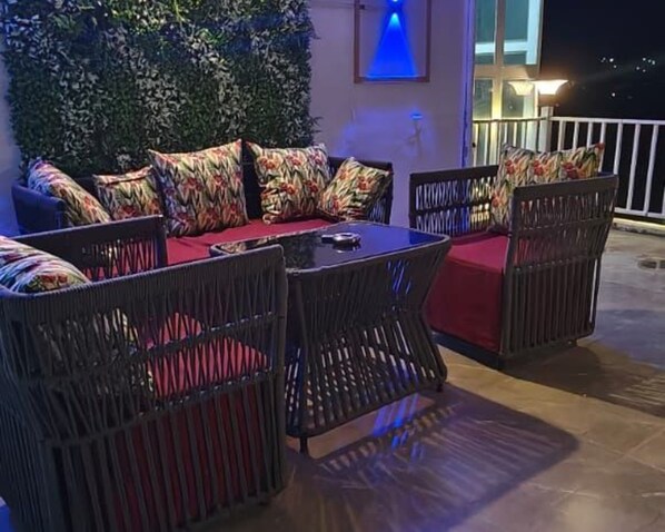 Un espacio al aire libre con mobiliario de mimbre y cojines rojos, una mesa y una pared iluminada en azul.