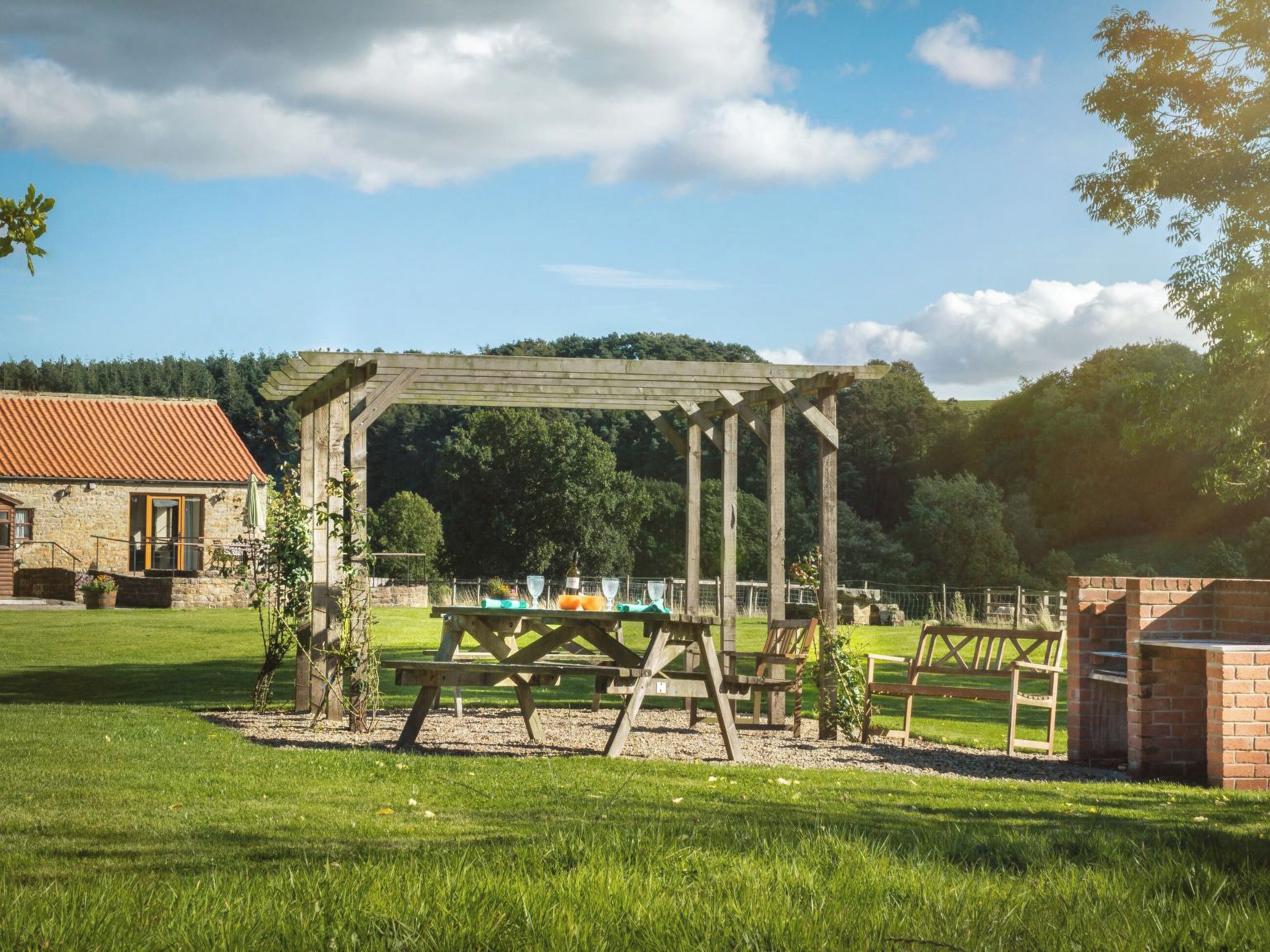 A wooden pergola with a picnic table and benches in a grassy area.