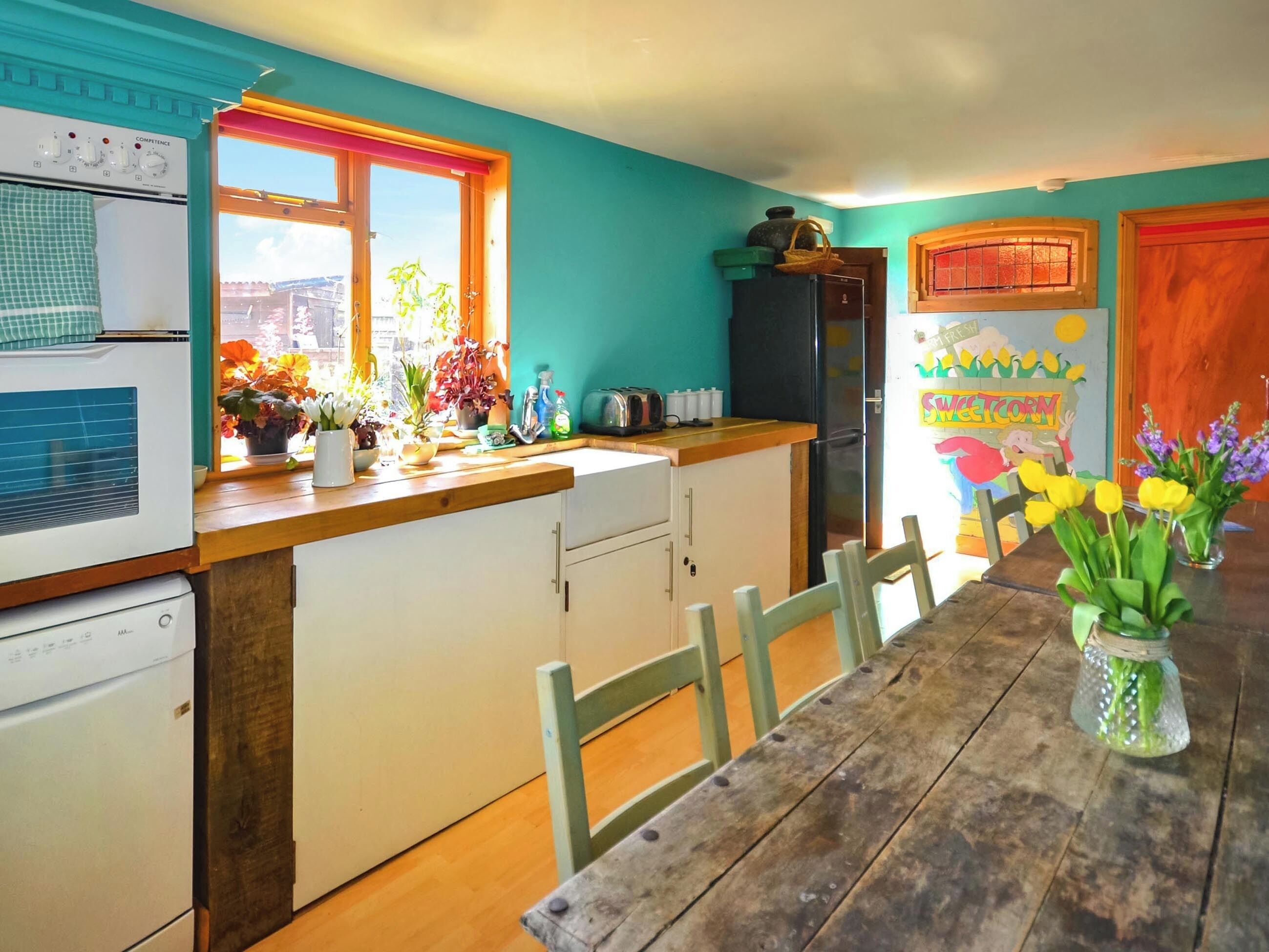 Une cuisine avec une table en bois, des chaises, un réfrigérateur, une cuisinière et une fenêtre ornée de plantes.