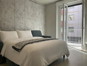 Ein modernes Schlafzimmer mit einem großen Bett, einem dunklen Sessel und Blick auf den Balkon.