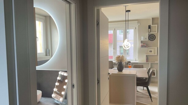 Ein modernes Interieur mit Schminkspiegel, Frisiertisch mit Beleuchtung, Blumen in einer Vase und einem Essbereich mit Stühlen und Tisch.