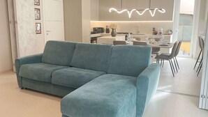 Ein modernes Wohnzimmer mit einem salbeengrünen Modulsystem, einem Essbereich mit Tisch und Stühlen und einer Küche im Hintergrund.