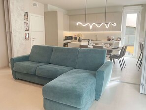 Un salon moderne avec un canapé d’angle turquoise, un coin repas avec une table et des chaises, et une cuisine en arrière-plan.