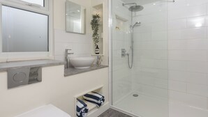 A modern bathroom with a glass shower enclosure, a white sink, and a built-in shelf with toiletries. - Little Dunley Cottages - Hazel (Bovey Tracey)
