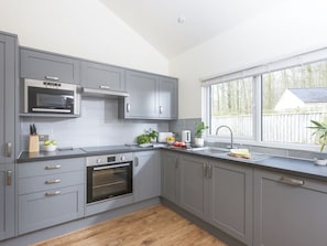 A modern kitchen with grey cabinets, a built-in oven, a microwave, and a window with a view of the outside. - Little Dunley Cottages - Maple (Bovey Tracey)