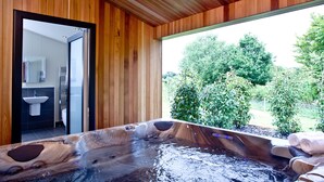 Un bain à remous avec un intérieur en bois, une vue sur une salle de bain avec un lavabo et un miroir, et un espace extérieur naturel agrémenté d’arbres.