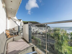 Ein Balkon mit Korbmöbeln, einer Glastür und Blick auf den Ozean sowie eine Küstenstadt.