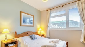 Un lit bien fait, avec du linge de lit blanc et une serviette pliée, une table de chevet en bois avec une lampe, un tableau encadré au mur et une fenêtre avec des rideaux.