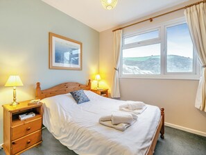 A neatly made bed with white linens and a folded towel, a wooden nightstand with a lamp, a framed picture on the wall, and a window with curtains. - A first floor apartment with sea views from the balcony. (Seaton)