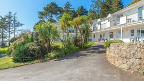Un bâtiment à deux étages doté d’un balcon, entouré d’une végétation luxuriante et d’un mur de pierre.