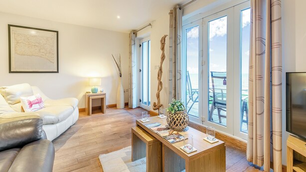 A modern living room with a sofa, coffee table, and a TV. Large windows offer a view of the sea. - 1 bedroom accommodation in Seaton (Seaton)