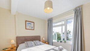 Un lit bien fait, avec des oreillers, une lampe de chevet, une table de nuit, une fenêtre avec des rideaux et une petite plante en pot posée sur le rebord de la fenêtre.